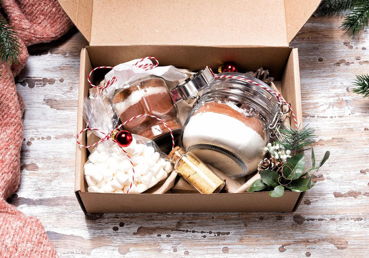 A festive gift box with jars of layered cocoa mix, mini marshmallows, and a small vial of golden glitter.