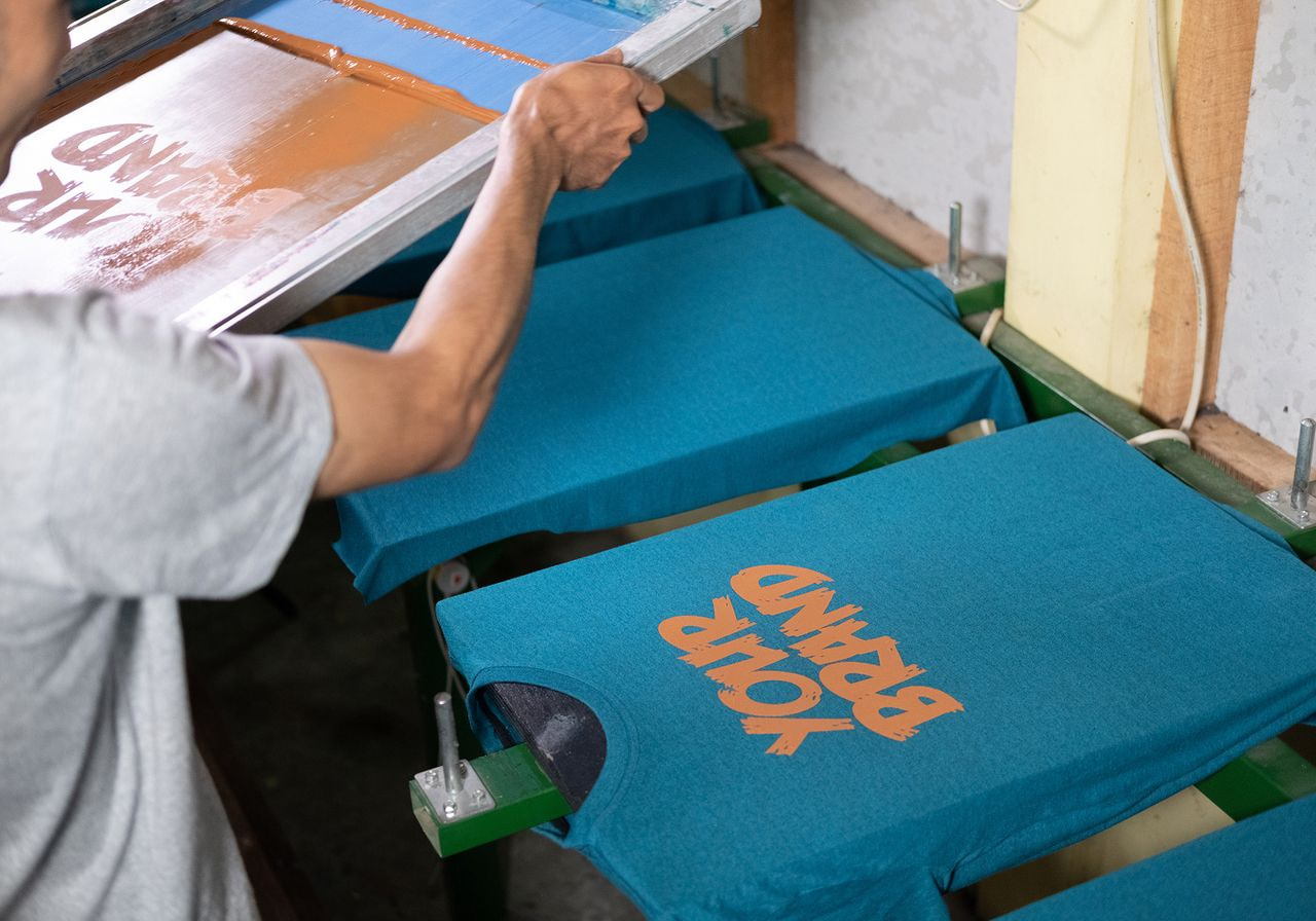 Man screen printing 'Your Brand' in orange ink onto teal t-shirts stretched on a press.