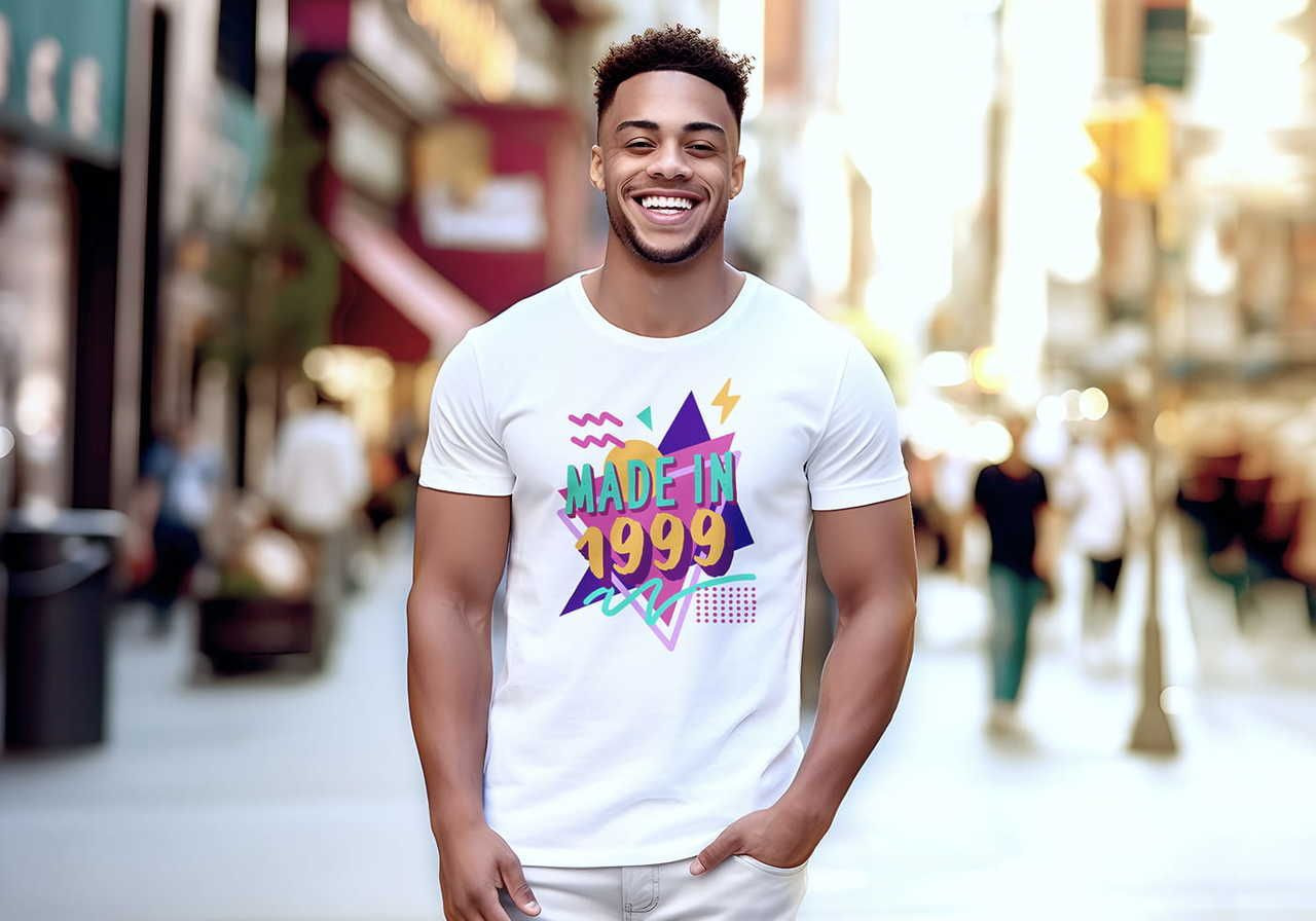 A smiling man in a white shirt with a colorful "Made in 1999" design stands on a sunny city street.