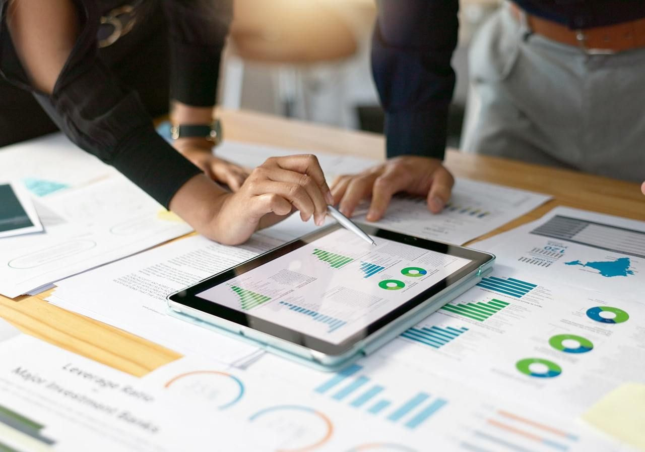 Two people analyze charts and graphs on a tablet and printed sheets spread across a desk.
