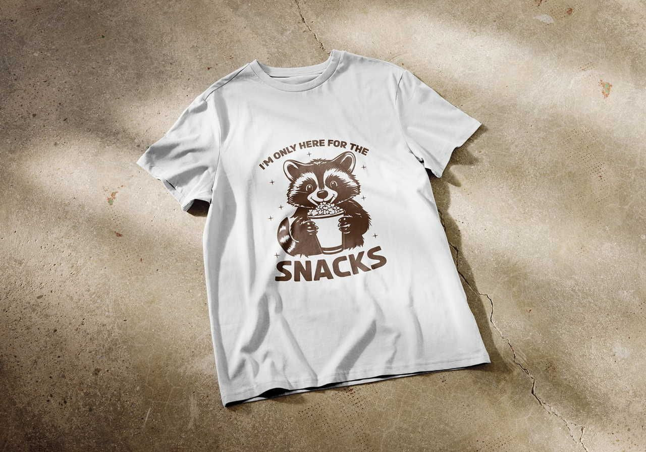 A white t-shirt on a concrete floor, showing a cute raccoon eating popcorn. Text reads "I’m only here for the snacks".