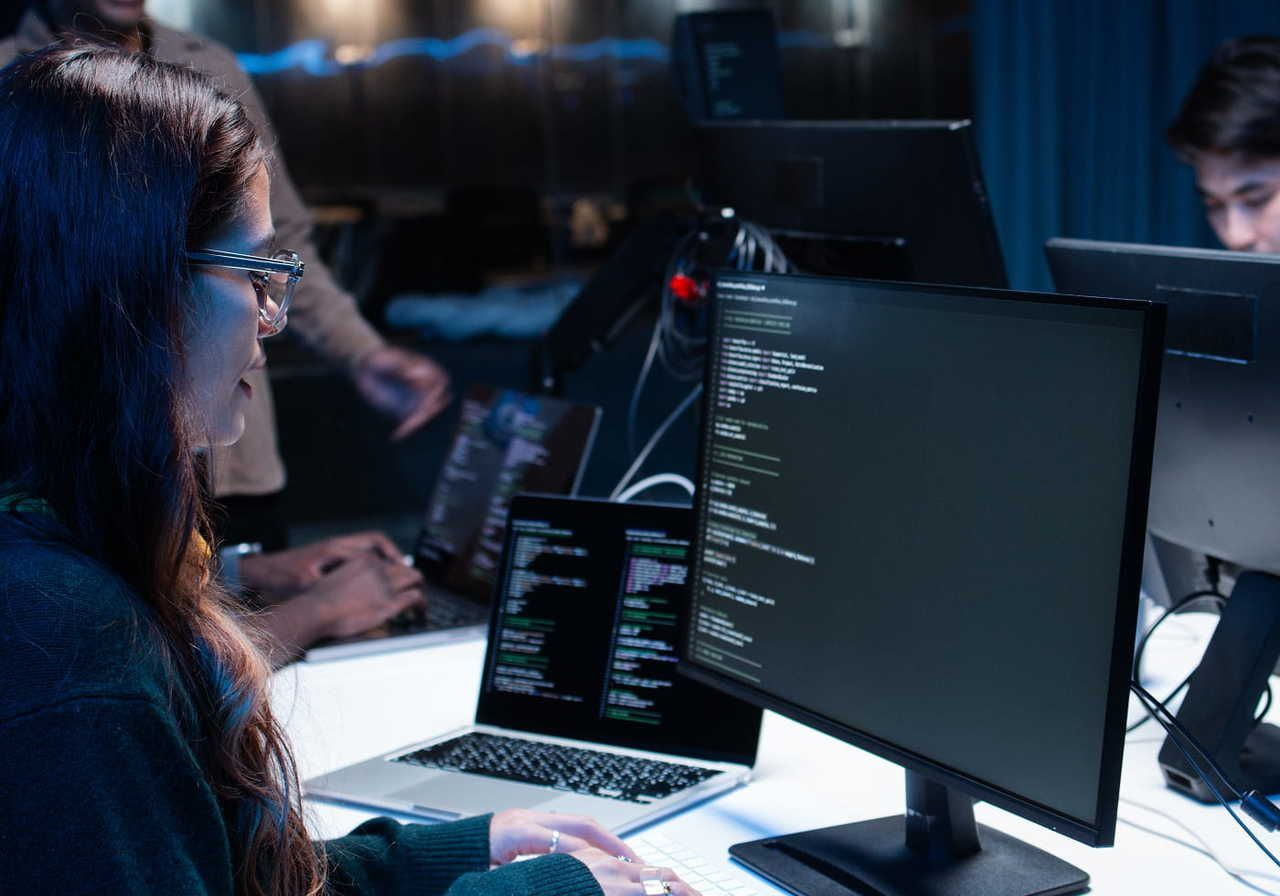 A woman in glasses is focused on coding at a computer. The room is dimly lit, and multiple screens display lines of code.