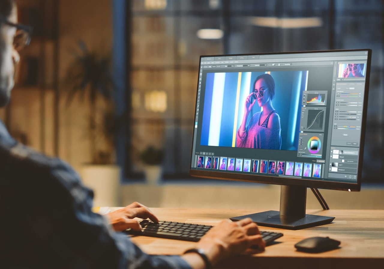 A man working on a computer with a monitor displaying an image editing software, showing a photo of a woman wearing sunglasses.