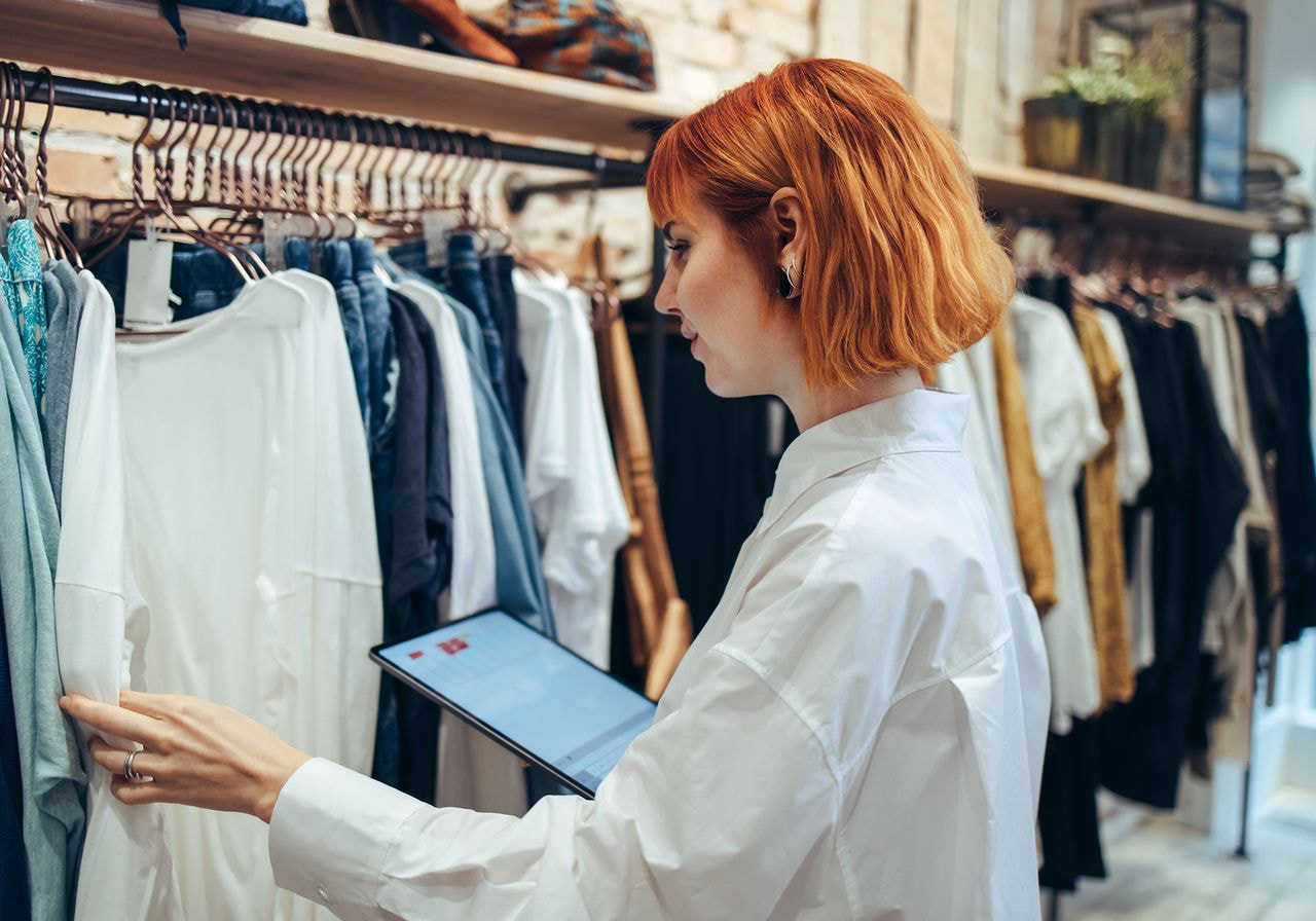 Woman with red hair checking clothing on a rack while holding a tablet in a store.