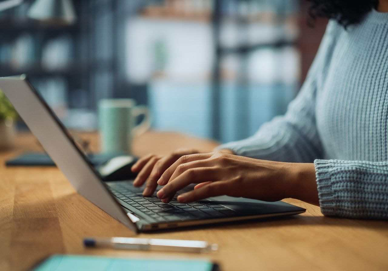Close-up of hands typing on a laptop at a wooden desk with a mug and pen nearby.