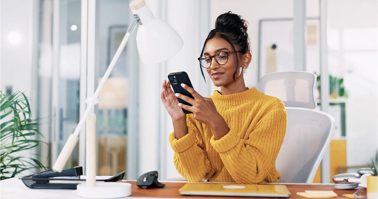 A smiling woman is sitting at a desk and looking at her mobile phone.