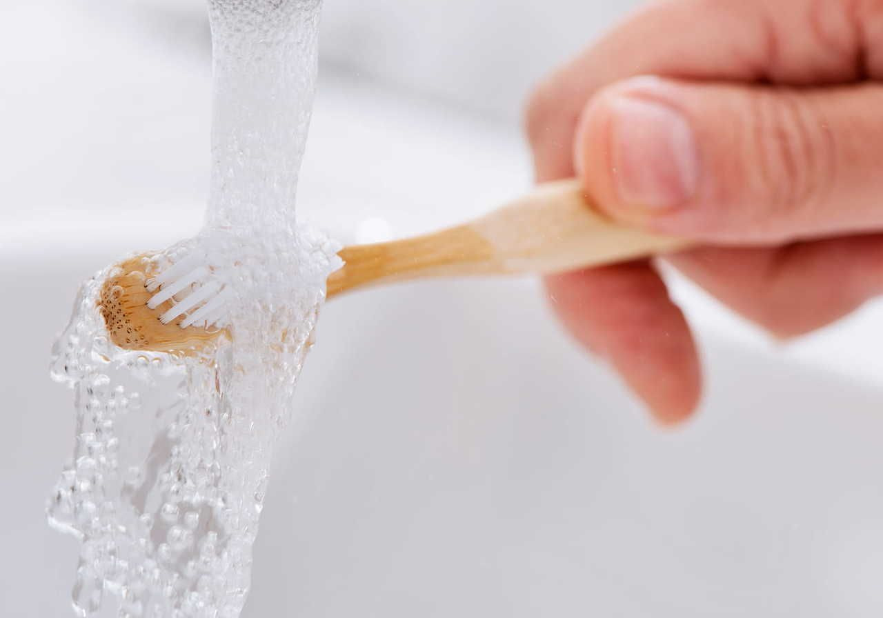 A hand rinses an eco-friendly bamboo toothbrush under a stream of water in a sink. The bristles are wet and glistening.