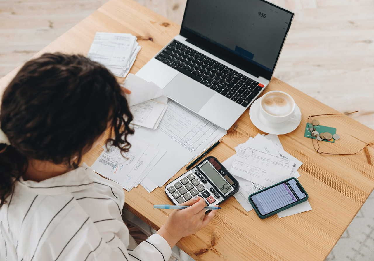 A woman using a calculator and a laptop while organizing paperwork on a wooden table.