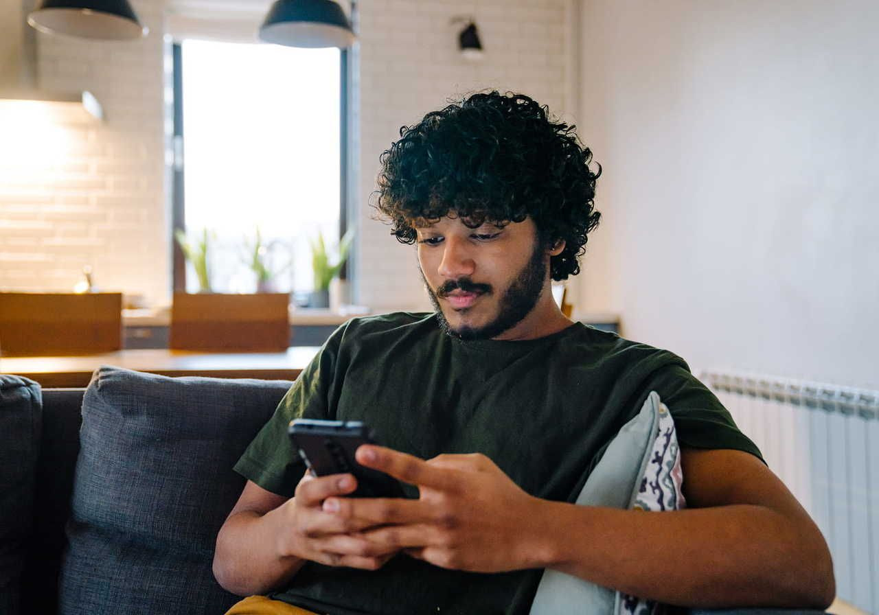 A young man with curly hair sitting on a couch, focused on his smartphone, searching for how to add products to TikTok Shop.