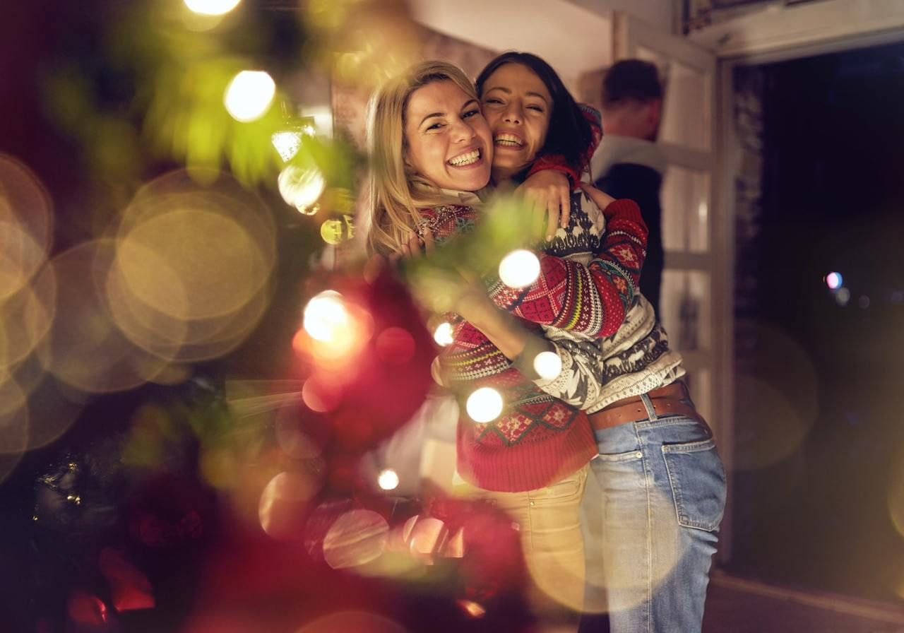 Two women embrace joyfully in front of a decorated Christmas tree, radiating warmth and holiday spirit.
