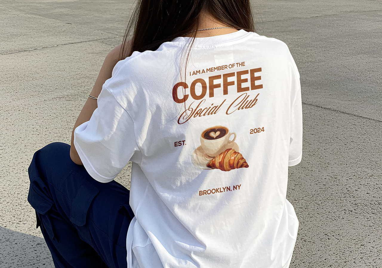A woman in a white t-shirt with "Coffee Social Club" and an illustration of a coffee cup and croissant on the back.