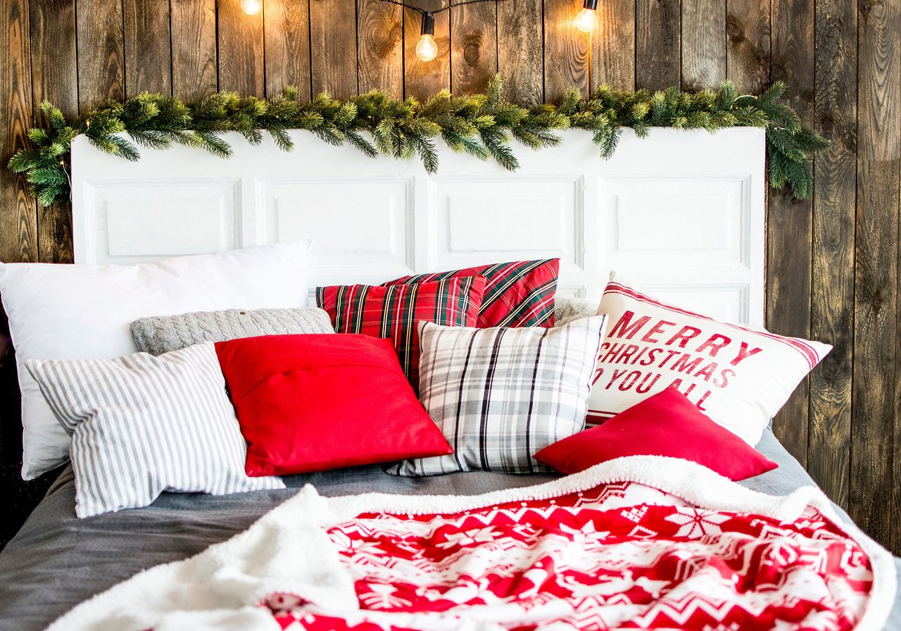 A cozy bed styled for the holidays with plaid, red, and “Merry Christmas” throw pillows, a red Nordic-pattern sherpa blanket, and a pine garland with string lights above the headboard.