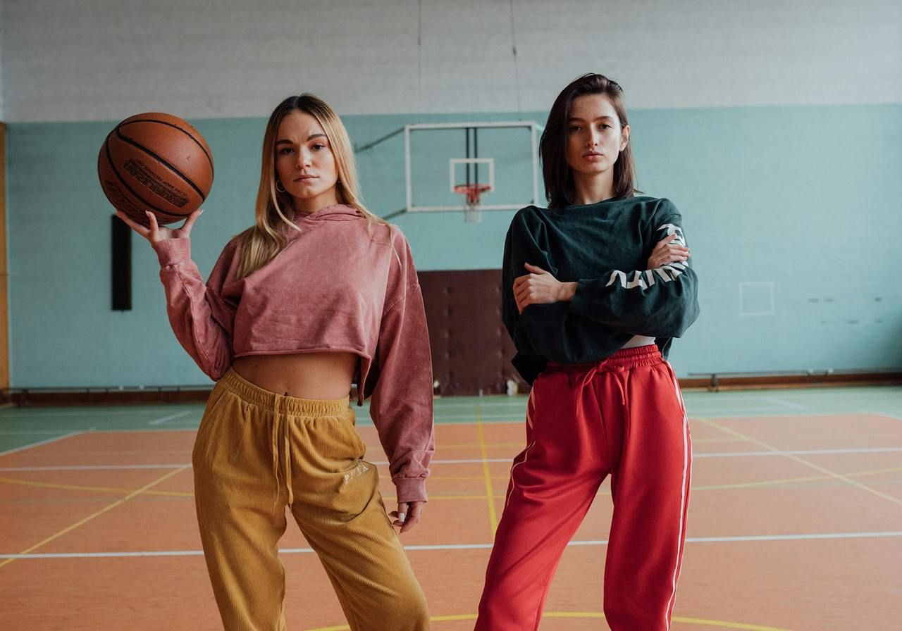 Two women dressed in hoodies and sports bottoms are playing basketball.
