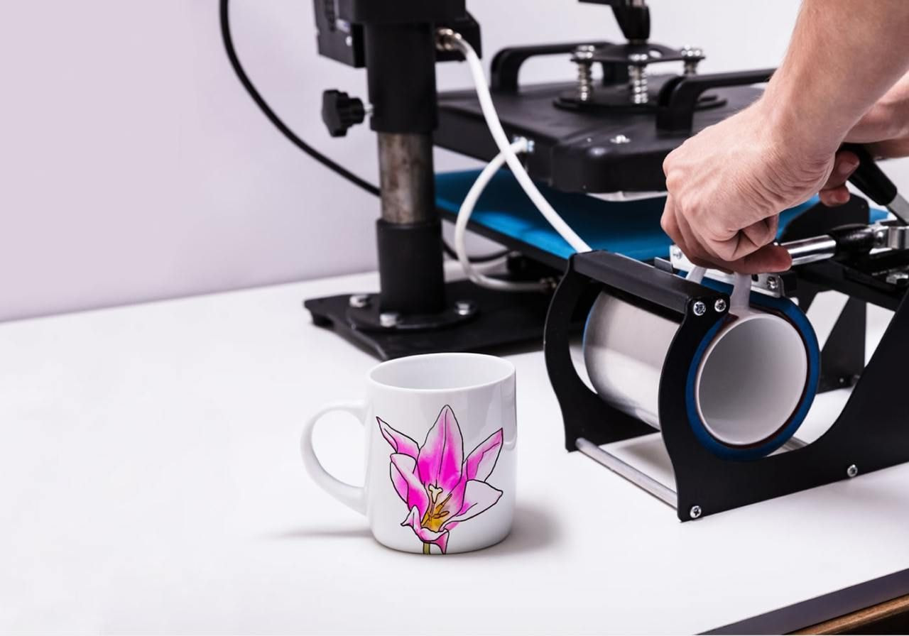 A person operates a printing machine to customize a mug with a design.