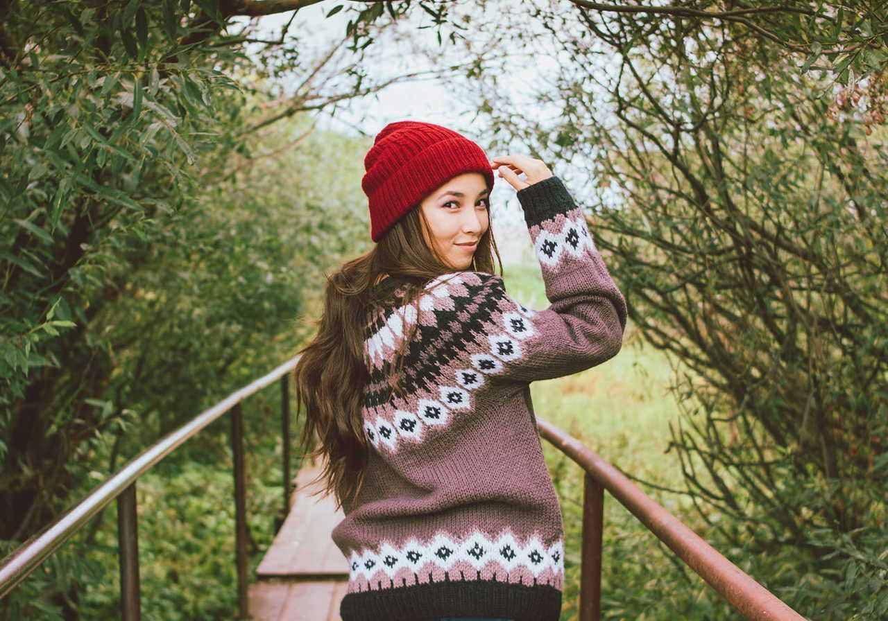 Young woman in a patterned knit sweater and red beanie looks back with a smile, standing on a narrow bridge surrounded by trees.