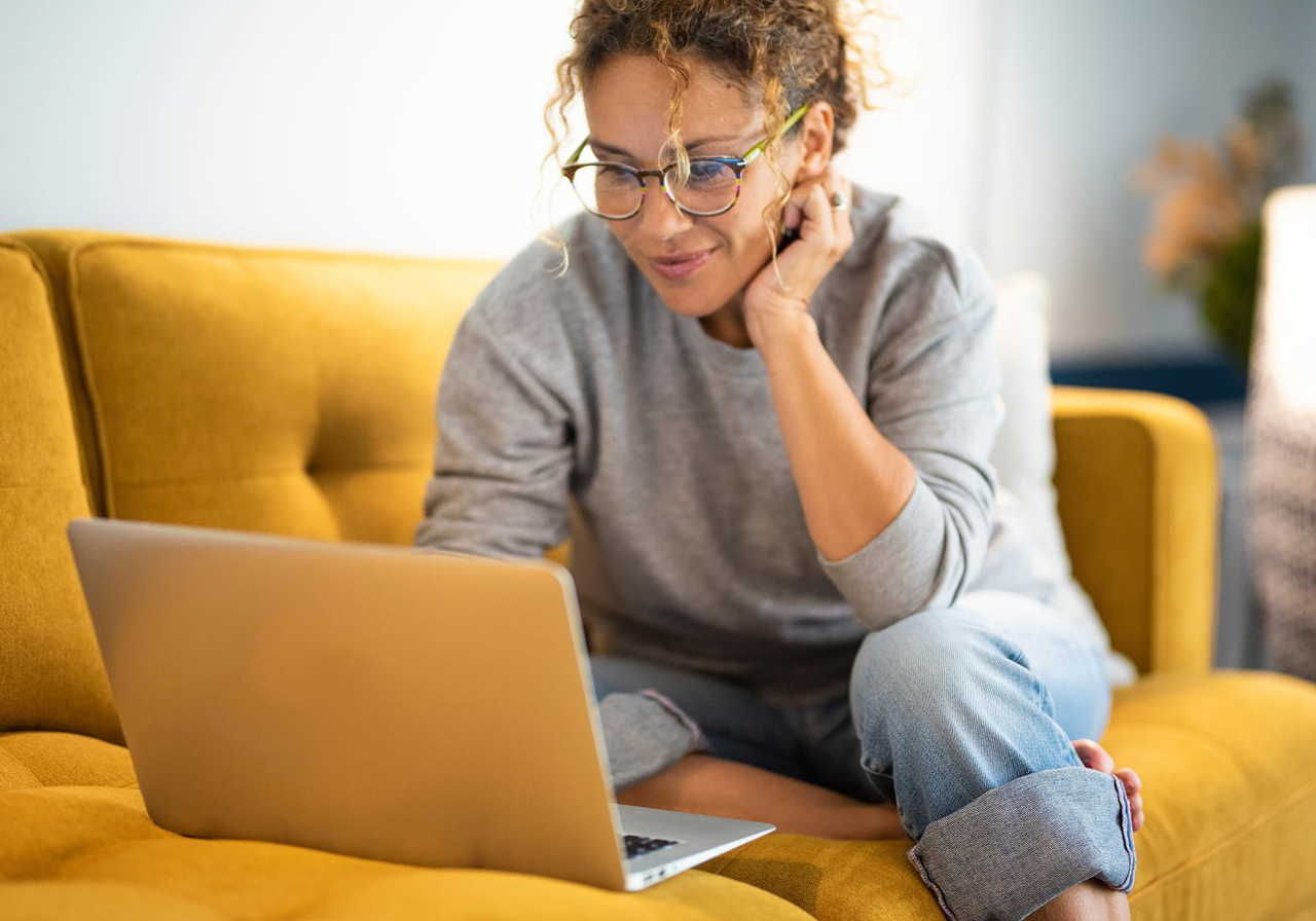 A smiling woman sitting on a yellow couch, looking at a laptop, reading a comparison Shopify vs Etsy. She is wearing glasses and has curly hair.