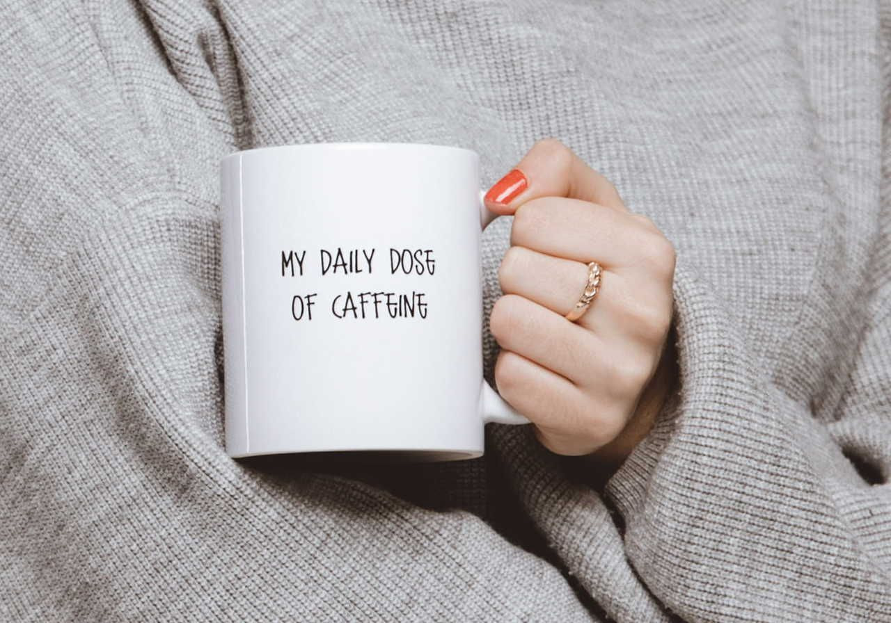 A person holding a white mug with the text "MY DAILY DOSE OF CAFFEINE" on it, wearing a gray sweater.