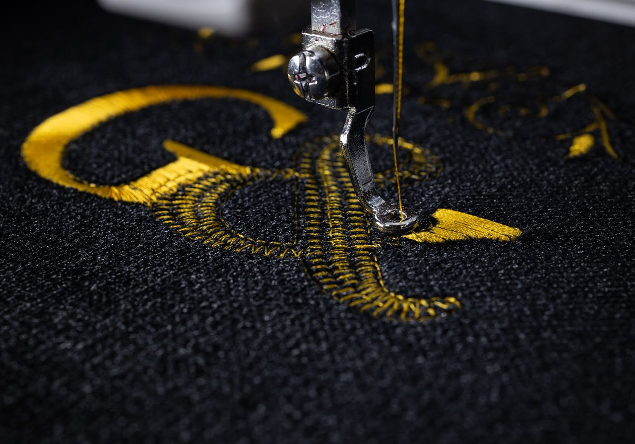 An embroidery machine needle stitches a gold thread design onto black fabric.