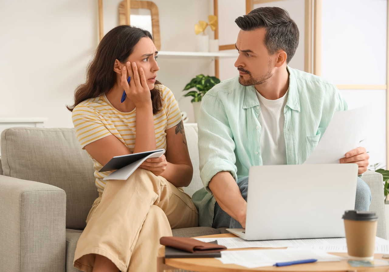 A man and a woman sitting on a couch, engaged in a serious discussion, with a laptop and papers in front of them.