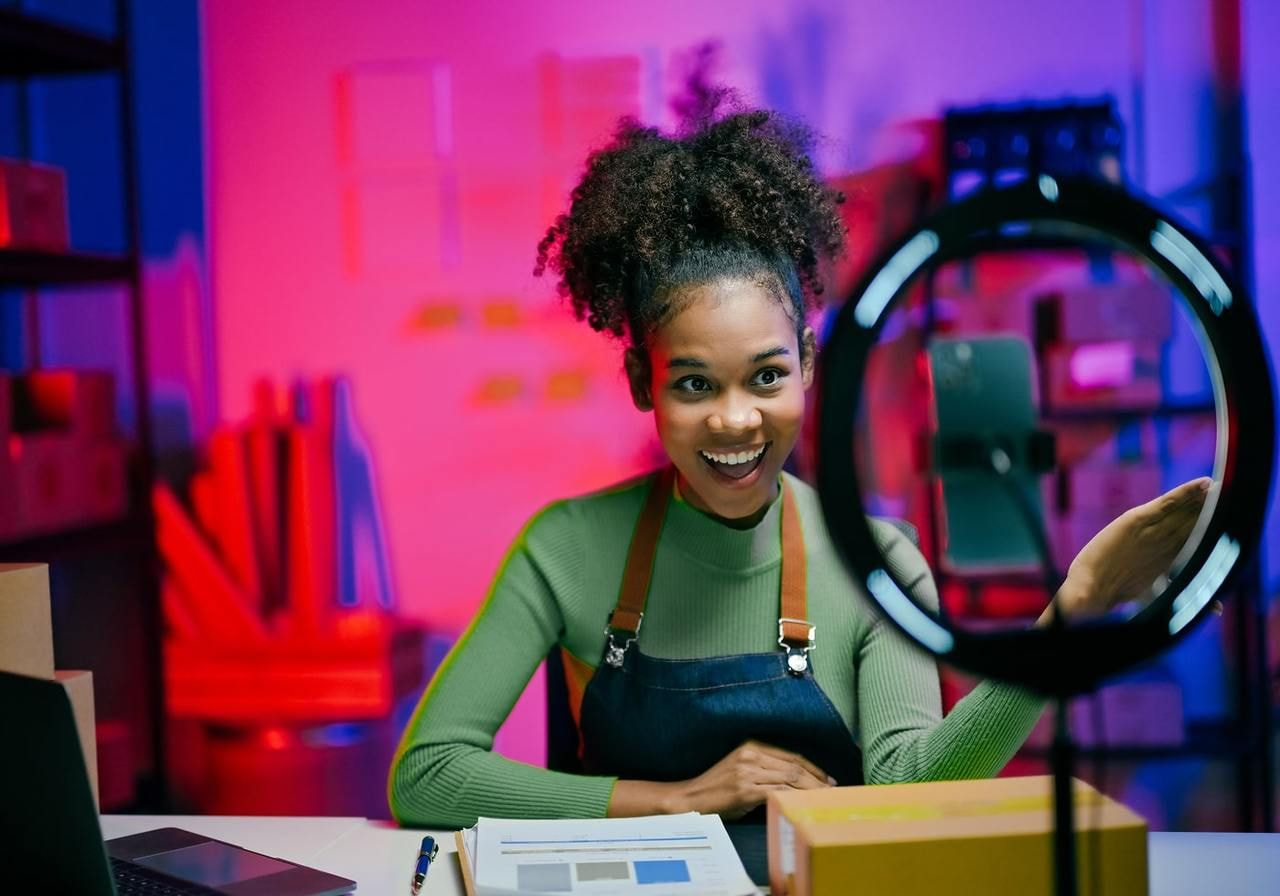 A woman wearing a green sweater and an apron waves joyfully at a smartphone on a tripod.