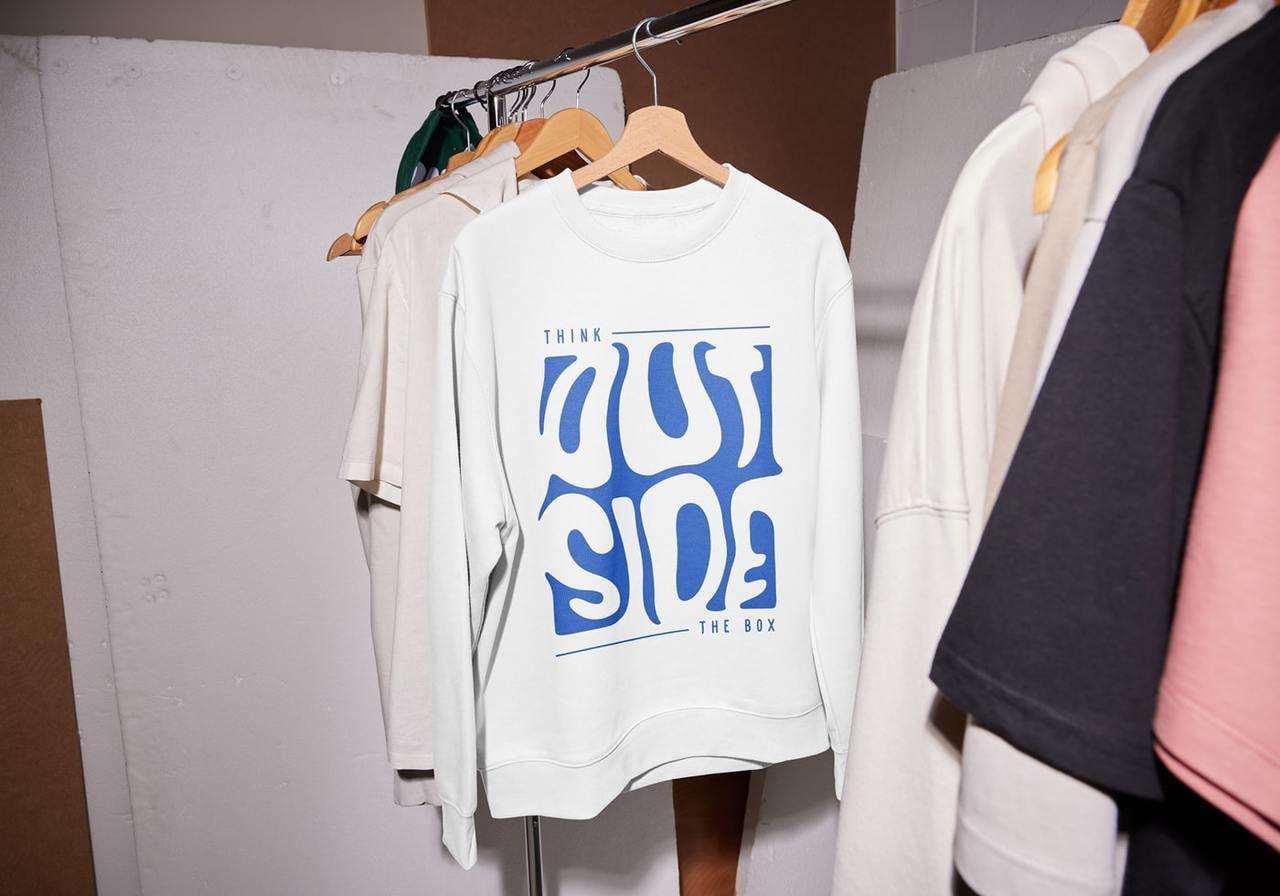 A white sweatshirt hangs on a rack among other clothes. Its bold blue text says, "Think Outside the Box.”