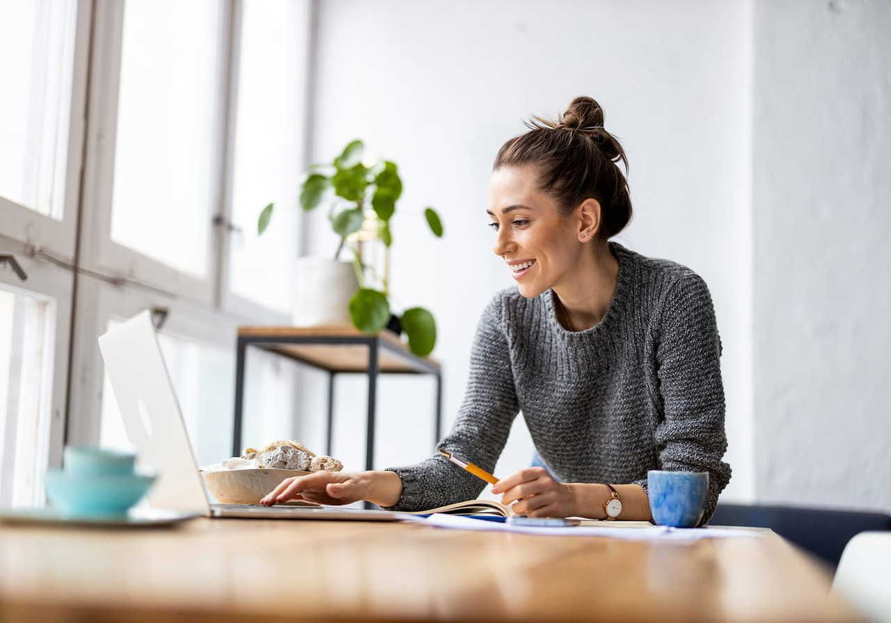 A woman smiling as she works on a laptop in a bright room. She holds a pencil, with a coffee mug and a plant nearby.