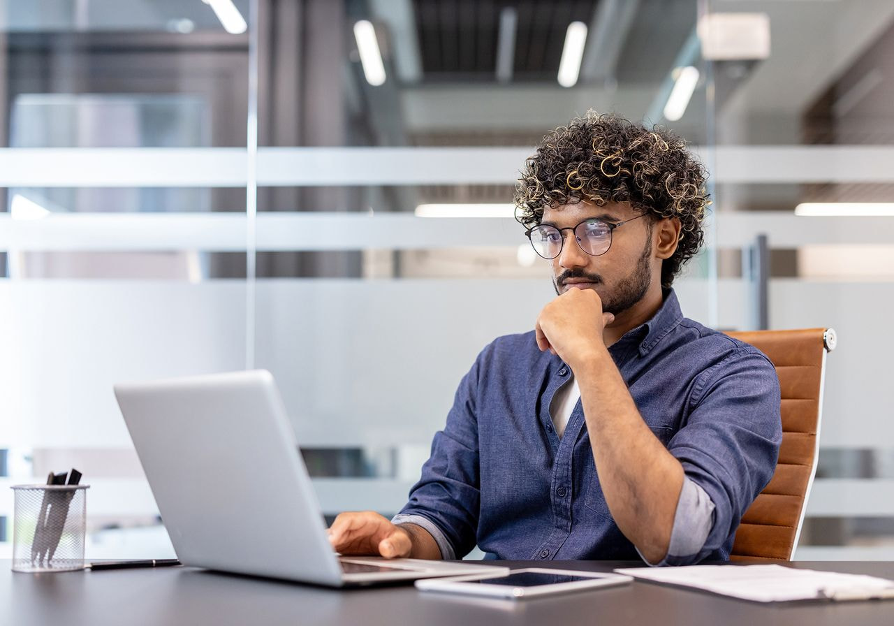 Man with curly hair thoughtfully looking at a laptop while working at a desk.
