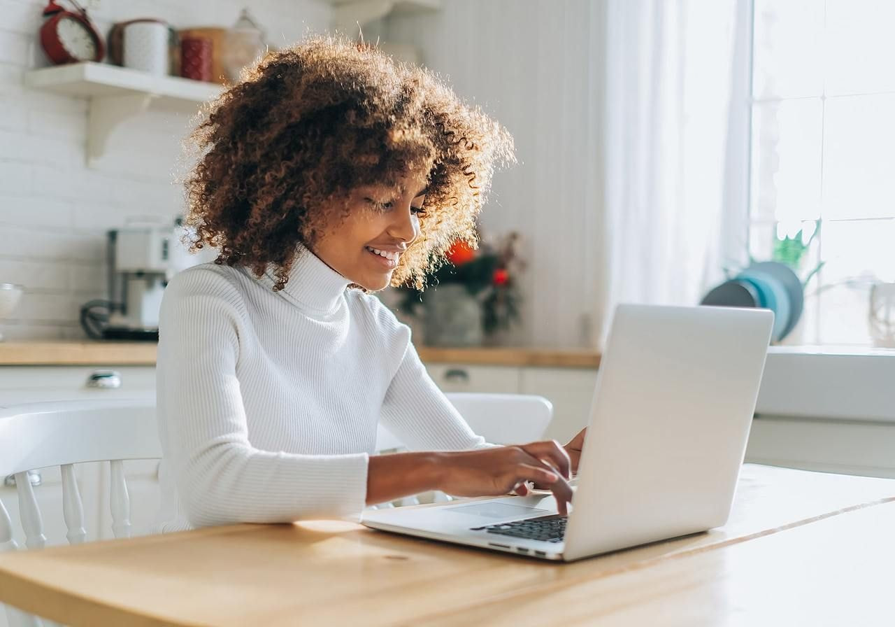A woman with curly hair sits at a table, focused on her laptop, surrounded by a cozy indoor setting.