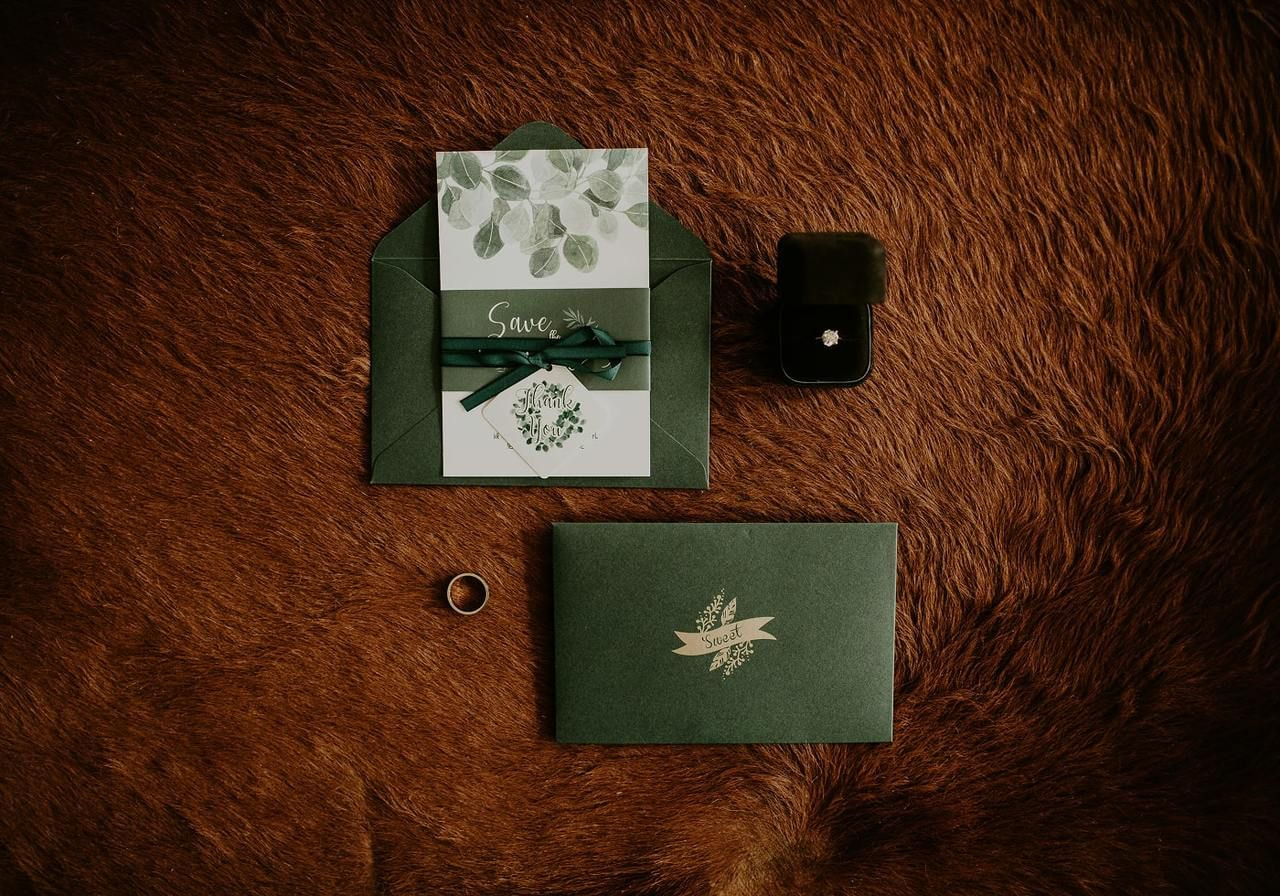 A green wedding invitation and envelope with a pen, placed on a soft brown fur surface.