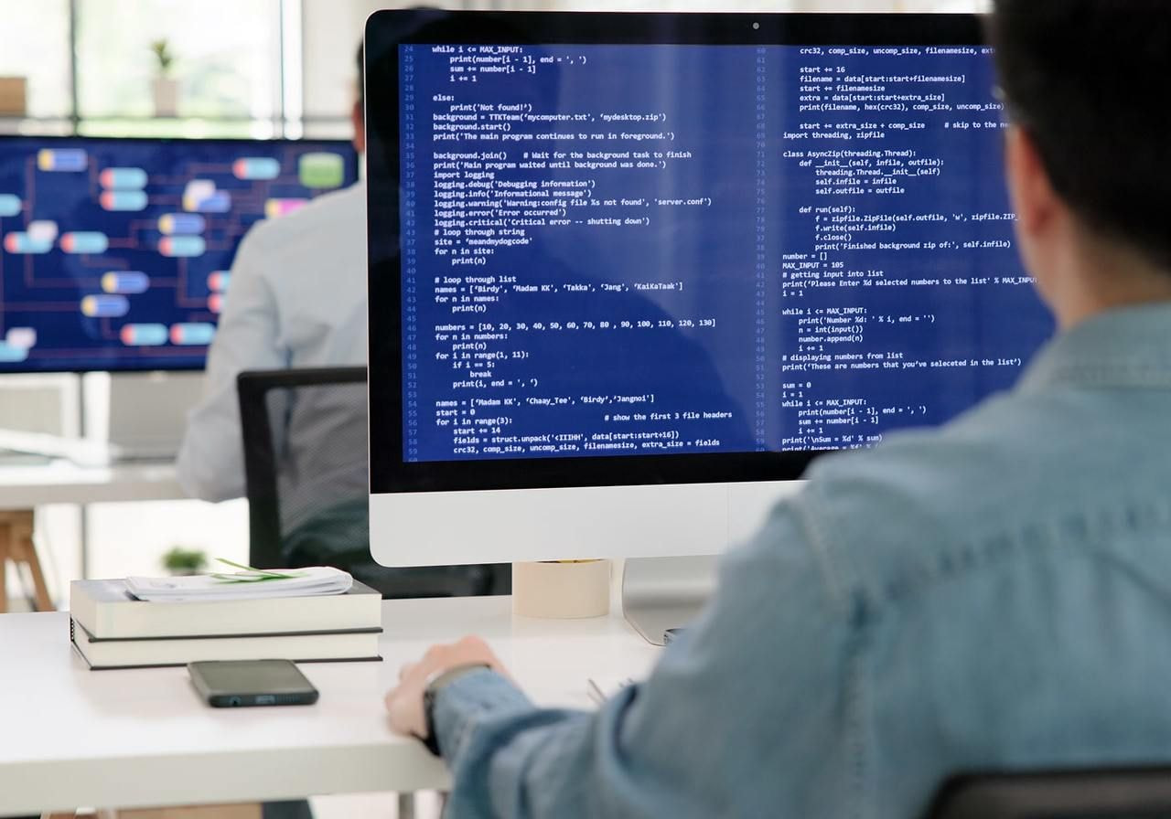 A person in a denim shirt works at a desk with a large monitor displaying code. Another screen with diagrams is in the background.