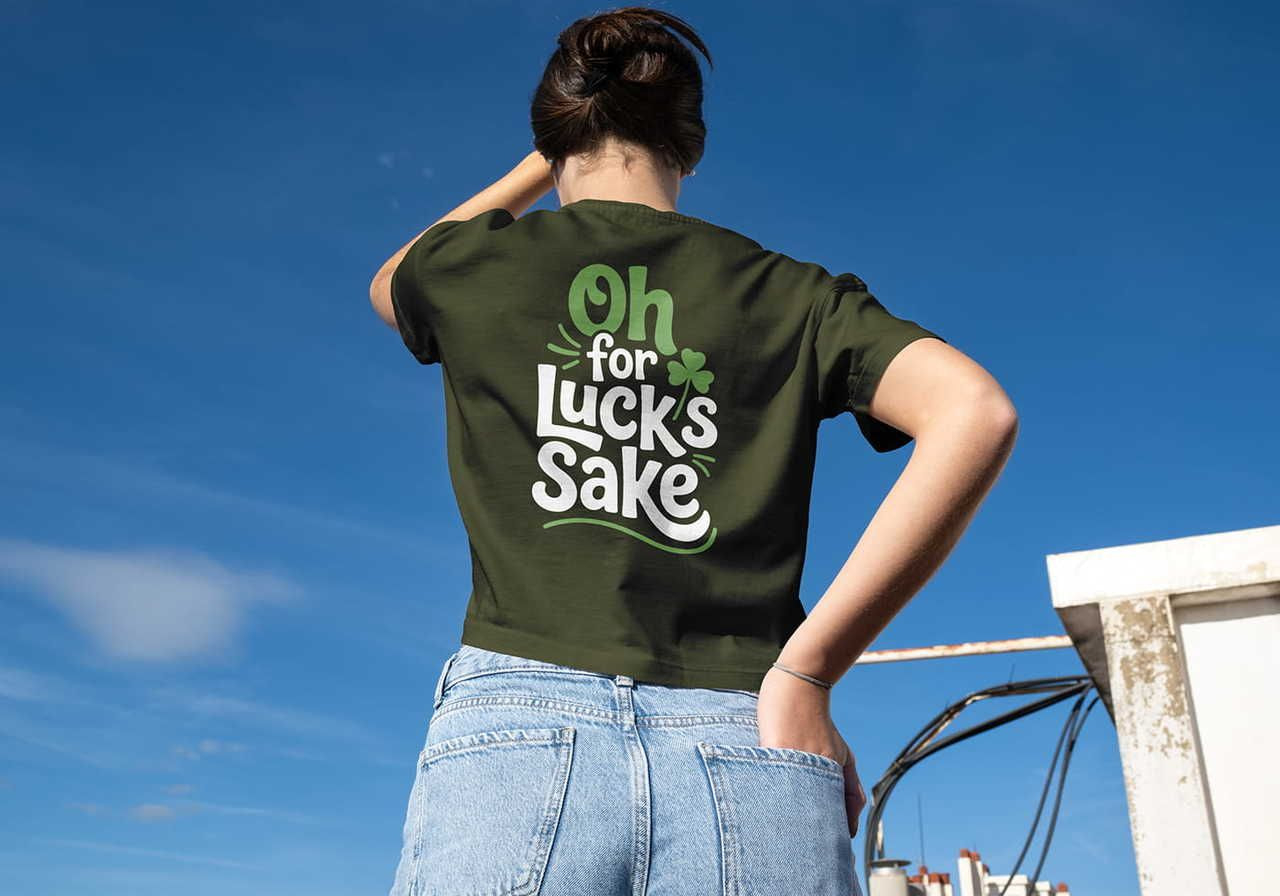 A person stands with their back to the camera under a blue sky, wearing a green "Oh for Luck's Sake" shirt and a small clover.