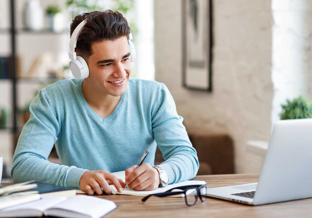 A man wearing headphones smiles as he writes in a notebook, sitting at a desk with an open laptop.