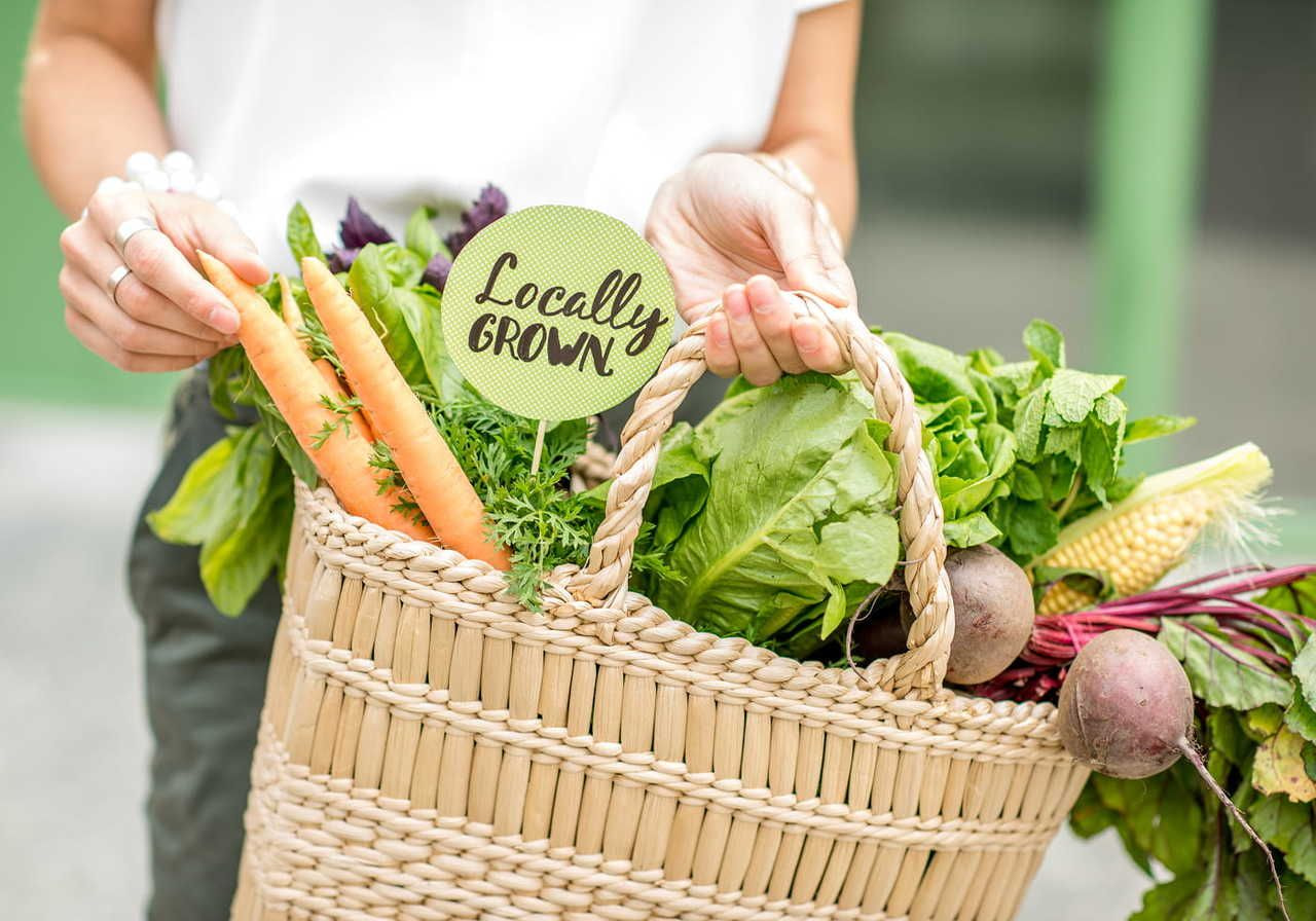 A person holding a woven basket filled with various fresh vegetables, including carrots, greens, and beets, with a sign that says "Locally GROWN".