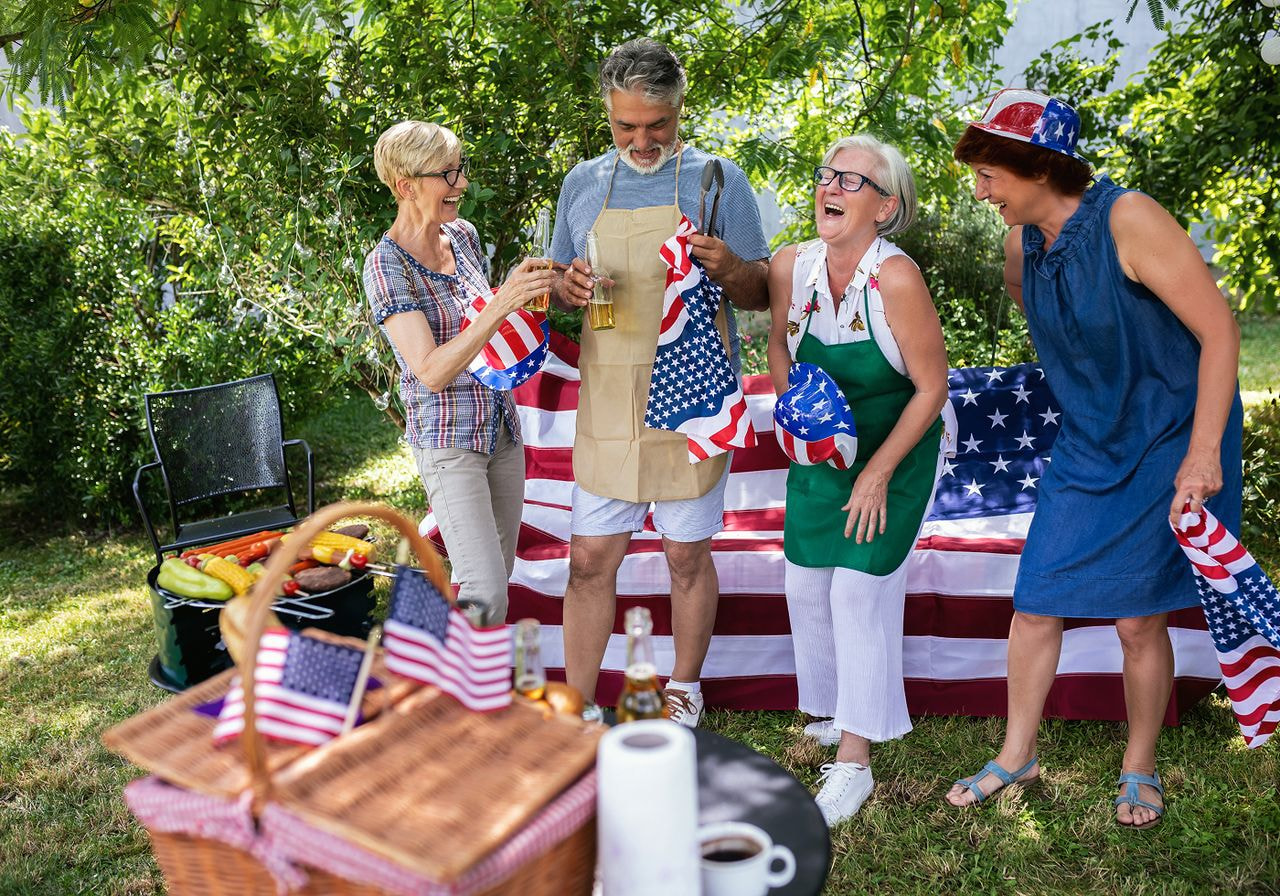 Four people enjoy a picnic in a yard adorned with American flags, surrounded by greenery and a festive atmosphere.