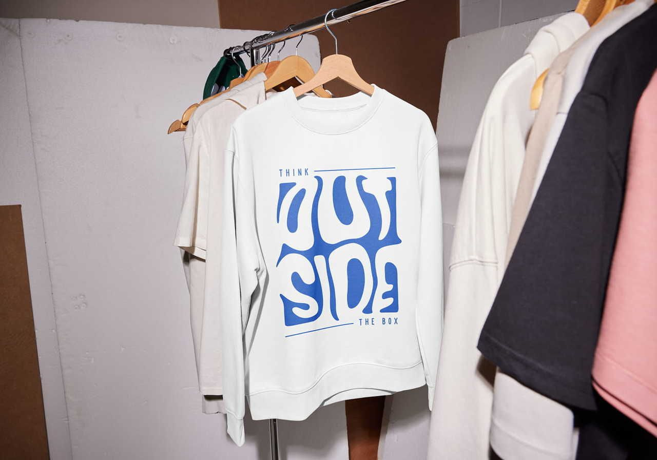 White sweatshirt hanging on a rack, featuring "Think Outside the Box" in stylized blue letters. Surrounded by other neutral-toned garments.