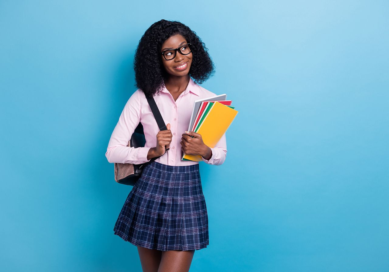 Smiling young woman with curly hair, wearing glasses and a pink shirt, holds books and a shoulder bag against a light blue background.