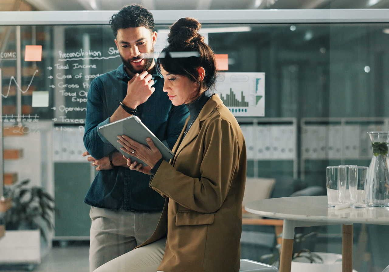 A man and a woman are engaged in a discussion in an office setting, with the woman holding a tablet and the man looking contemplative.