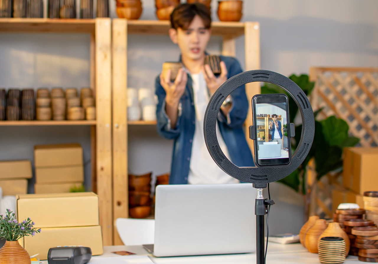 A man livestreams with a phone on a ring light, showcasing pottery. Shelves with wooden bowls and cardboard boxes are behind him.