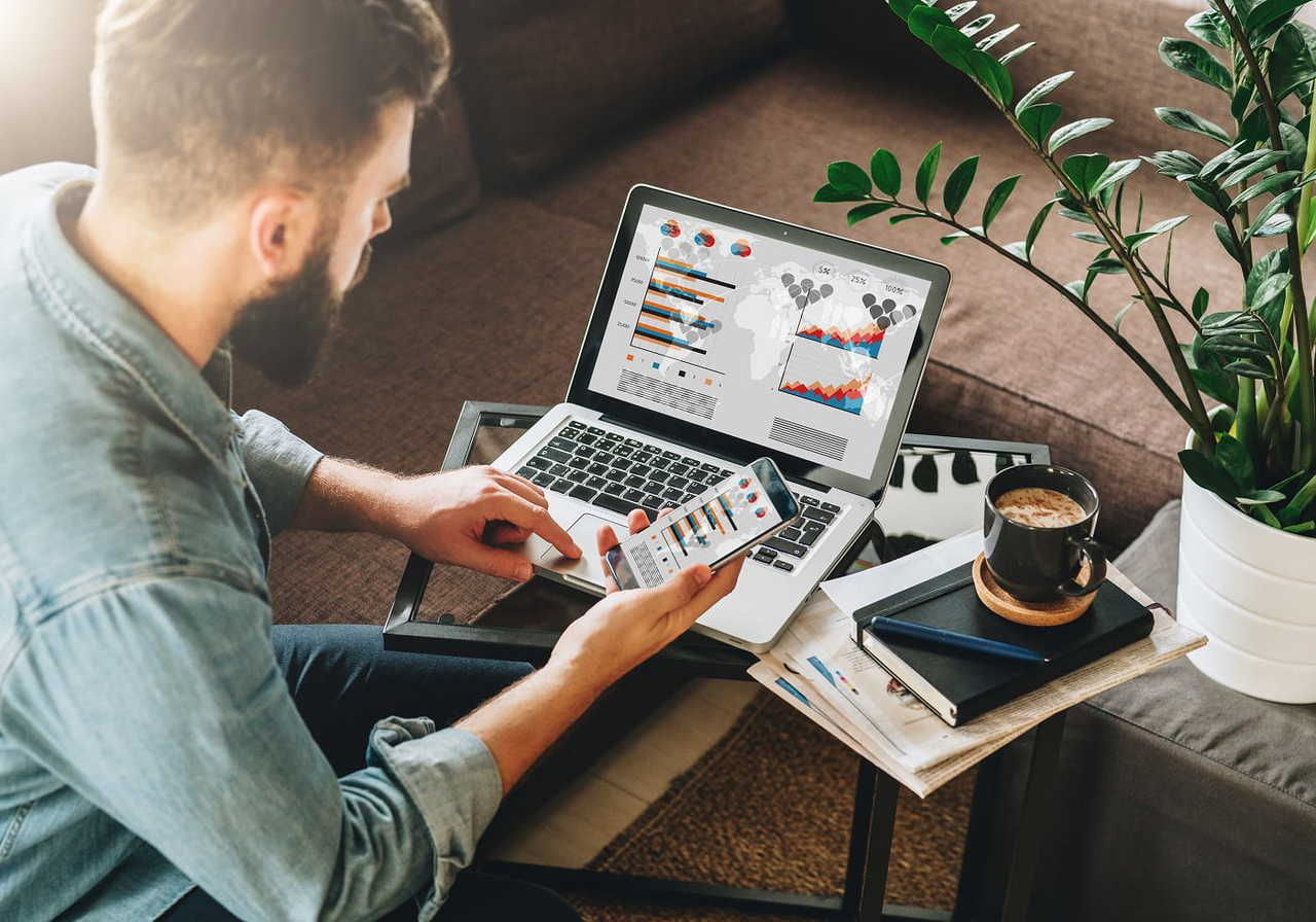 A man analyzing graphs on a laptop and smartphone at home, sitting on a sofa. Nearby, a coffee mug, notebook, and potted plant.