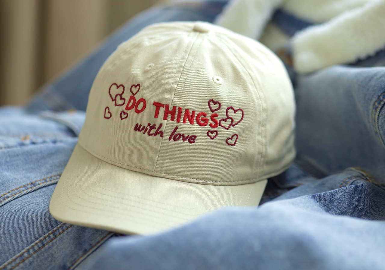 A white cap with “DO THINGS with love” and hearts embroidered, lying on top of denim fabric.
