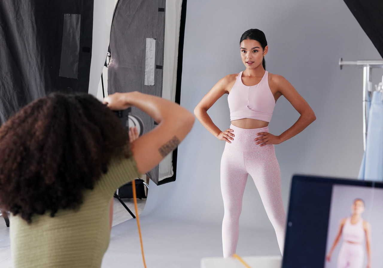 A fitness model poses in a pink athletic outfit while a photographer takes her picture in a studio setting.