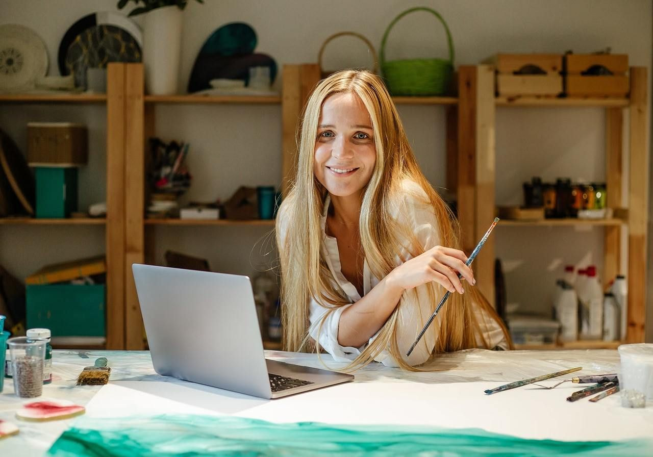 A smiling woman artist is sitting in front of a laptop, holding a brush.
