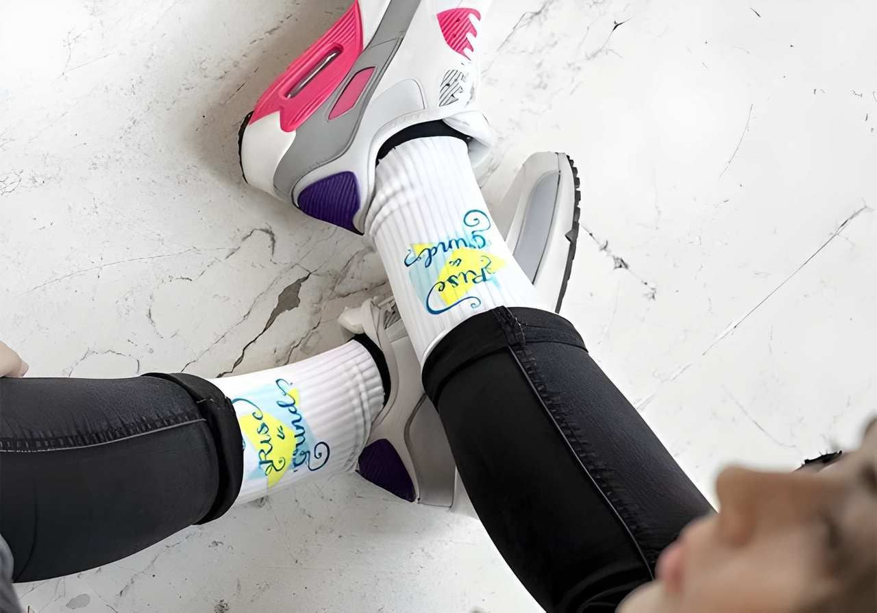 Woman wearing pink and gray sneakers with white socks featuring colorful cursive text. She is seated casually on a white marble floor.