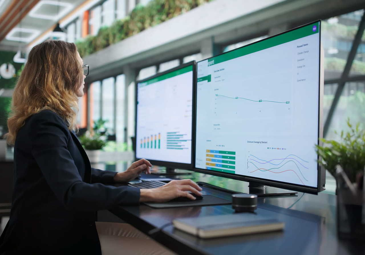 A woman sitting at a desk, using a computer with dual monitors displaying graphs and data analytics in a modern office environment.