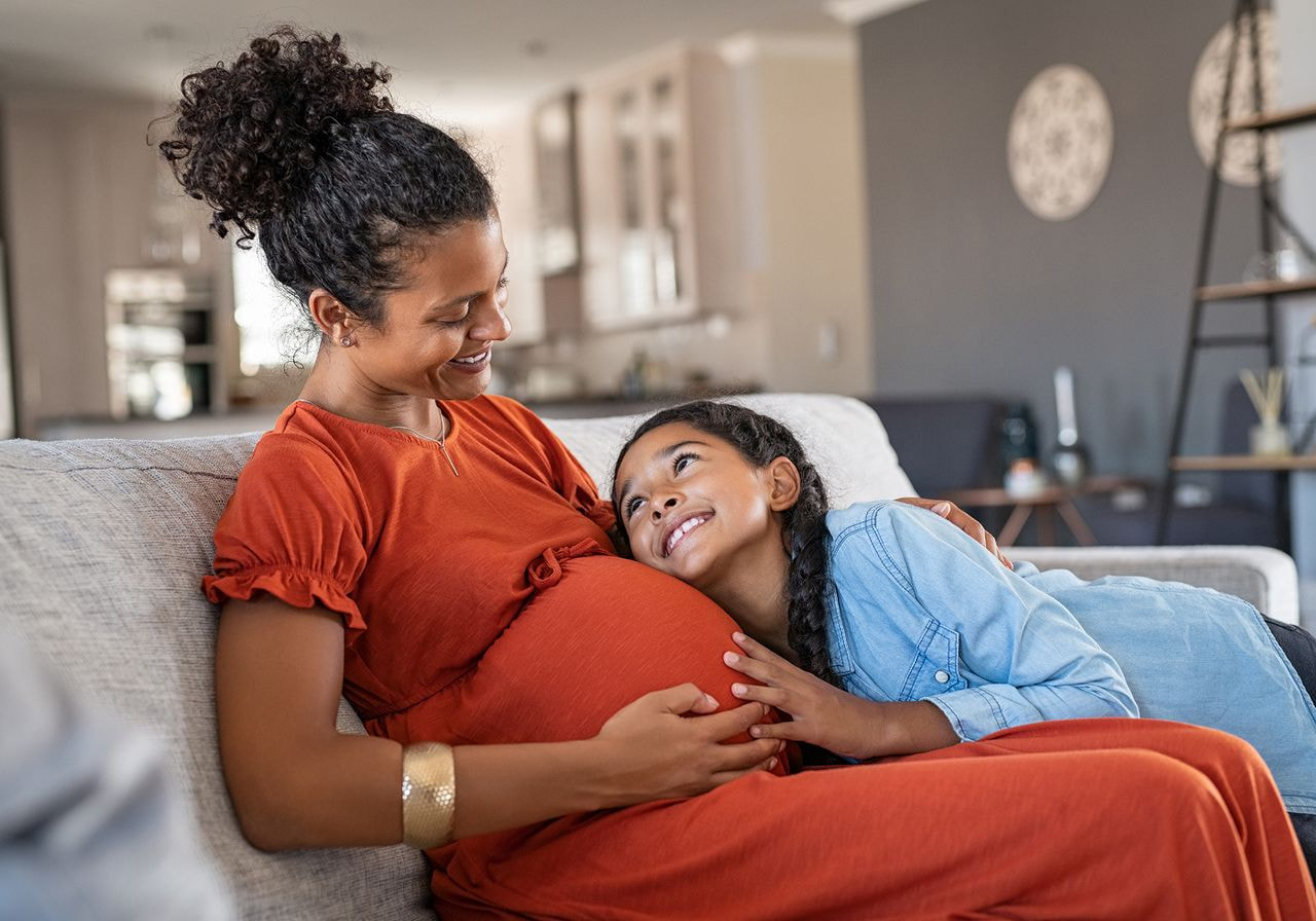A pregnant woman and her young daughter sit together on a couch, sharing a moment of warmth and connection.