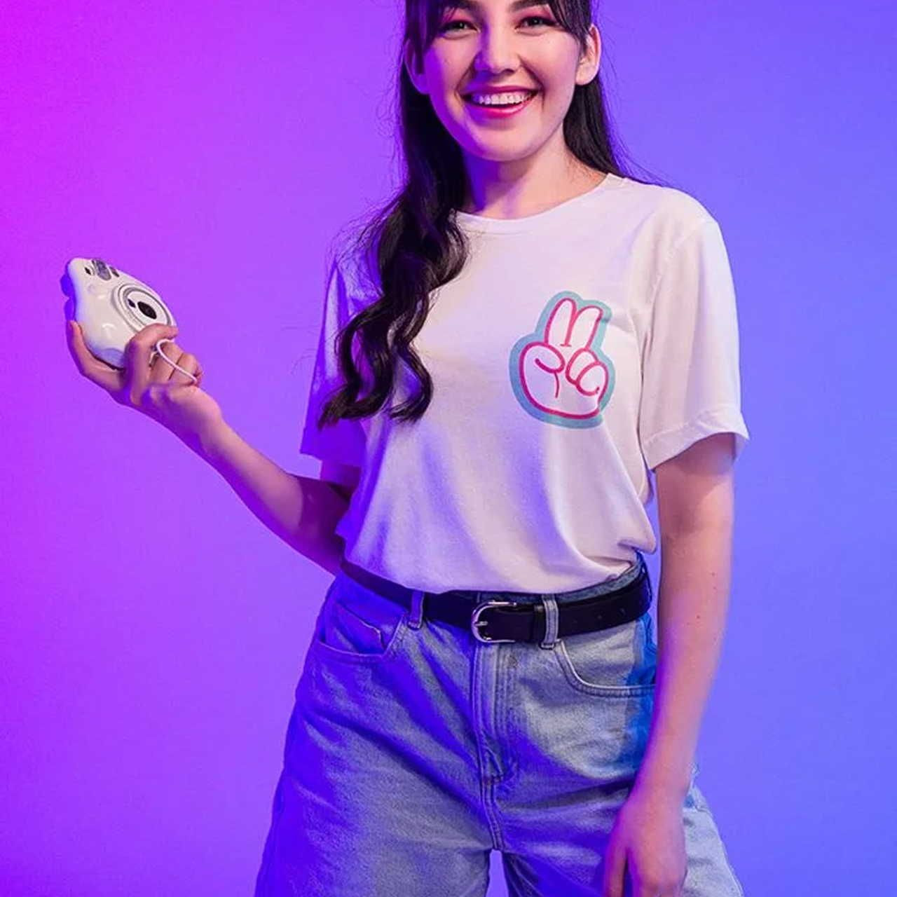 A smiling woman holding a gamepad, wearing a white Unisex Tri-Blend T-Shirt | Bella + Canvas 3413 with an illustrated hand piece sign.
