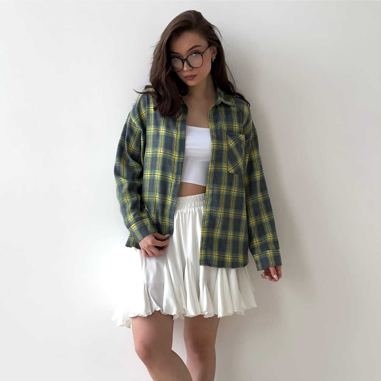 A young woman in round glasses wears a plaid flannel shirt over a white crop top and flowy white skirt.
