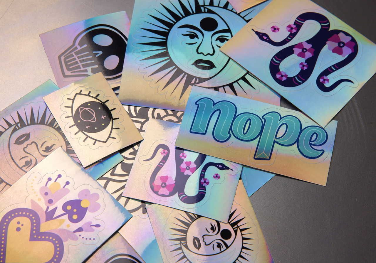 A collection of holographic stickers featuring a skull, sun-and-moon face, snakes, flowers, an eye, a heart, and the word "nope".