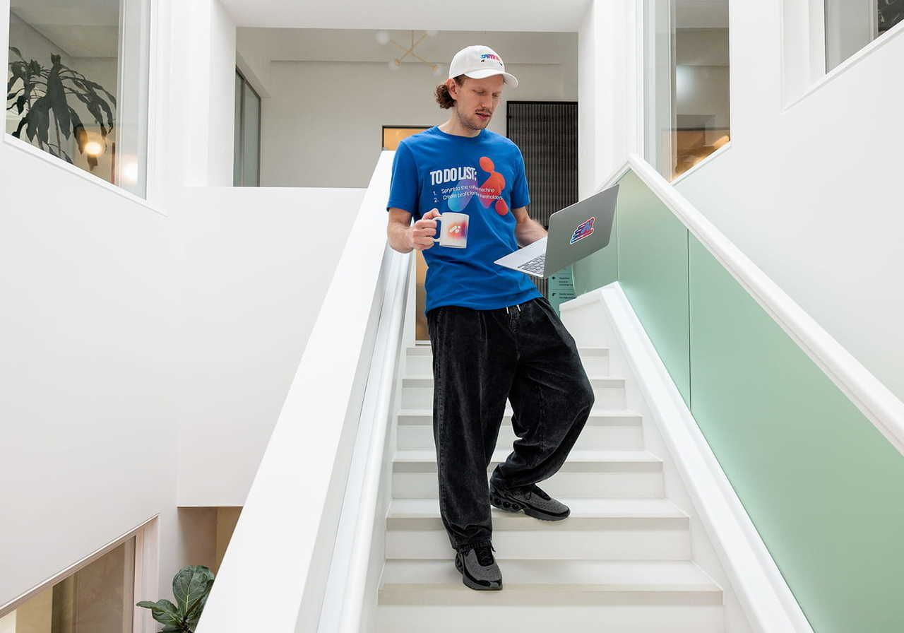 A young man wearing a blue t-shirt and black pants is walking downstairs while holding a laptop and a coffee mug.