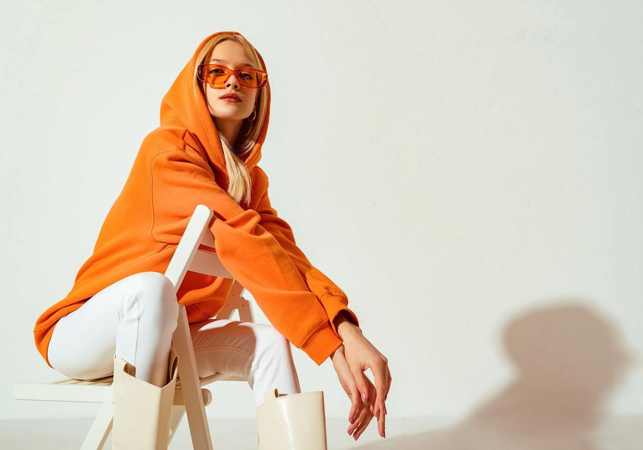 A woman in a vibrant orange hoodie and matching sunglasses sits on a white chair, wearing white pants and knee-high boots.